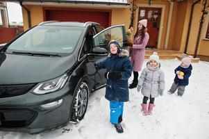 giovane donna con bambini tiene borse ecologiche e carica un'auto elettrica nel cortile di casa sua. foto