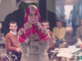 ritratto di giovane musulmano donna indossare hijab nel ufficio mentre guardare a telecamera. vicino su viso di Arabo attività commerciale donna coperto con foulard sorridente. riuscito arabo donna d'affari nel moderno ufficio. foto