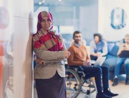 ritratto di giovane musulmano donna indossare hijab nel ufficio mentre guardare a telecamera. vicino su viso di Arabo attività commerciale donna coperto con foulard sorridente. riuscito arabo donna d'affari nel moderno ufficio. foto