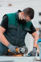 interno di una fabbrica di ingegneria dell'industria pesante con un lavoratore industriale che utilizza una smerigliatrice angolare e taglia un tubo di metallo. appaltatore in uniforme di sicurezza e elmetto per la produzione di strutture metalliche. foto