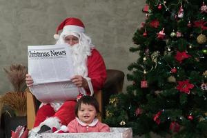 ritratto adorabile infanzia ragazza contento sorridente nel un' grande regalo scatola vicino il Natale albero. Grasso caucasico vecchio maturo uomo nel costume Santa Claus contento bianca barba seduta su il divano lettura giornali. foto