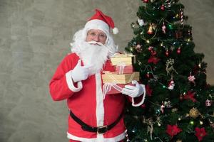 Grasso caucasico vecchio maturo uomo nel costume Santa Claus contento bianca barba sorridente Tenere regalo scatole preparazione decorazioni per Natale albero Festival celebrazione. allegro Natale contento nuovo anno concetto foto