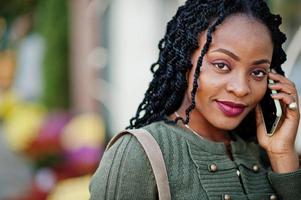 donne afroamericane alla moda alla moda in maglione verde e gonna nera poste in un caffè all'aperto con una tazza di caffè e parlare sul telefono cellulare. foto