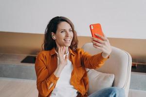 sorridente femmina blogger parlare su video chiamata su Telefono, saluto nel sociale reti, seduta nel poltrona foto
