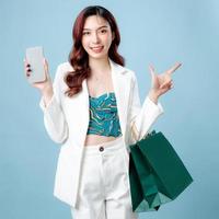 metà corpo di bellissimo asiatico attività commerciale donna indossare bianca completo da uomo puntamento per vuoto spazio Tenere shopping Borsa e mobile Telefono su isolato blu sfondo. vuoto schermo smartphone concetto. foto