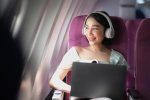 asiatico giovane donna indossare cuffie utilizzando il computer portatile seduta vicino finestre a primo classe su aereo durante volo, in viaggio e attività commerciale concetto foto