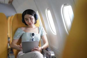 viaggio turismo con moderno tecnologia e aria voli concetto, donna seduta nel aereo con moderno digitale aggeggio e ricerca preferito musica elenco di riproduzione nel applicazione per ascoltando foto