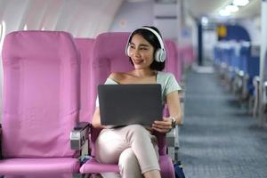 asiatico giovane donna indossare cuffie utilizzando il computer portatile seduta vicino finestre a primo classe su aereo durante volo, in viaggio e attività commerciale concetto foto