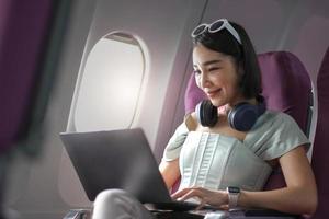 asiatico giovane donna utilizzando il computer portatile seduta vicino finestre a primo classe su aereo durante volo, viaggio e attività commerciale concetto foto