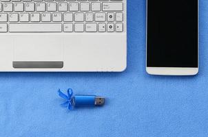 brillante blu USB veloce memoria carta con un' blu arco bugie su un' coperta di morbido e peloso leggero blu vello tessuto accanto per un' bianca il computer portatile e smartphone. classico femmina regalo design per un' memoria carta foto