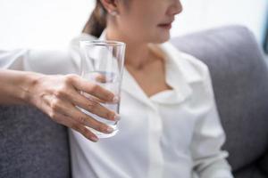 giovane asiatico donna Tenere bicchiere di puro minerale acqua acqua nel mani e seduta su confortevole divano a casa, sensazione rinfrescato energico nel mattina. foto