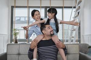 asiatico tailandese famiglia insieme, papà giochi e stuzzica con figlia e mamma di trasporto e Tenere ragazza su le spalle nel casa vivente camera, contento tempo libero volte, bello fine settimana, benessere domestico stile di vita. foto