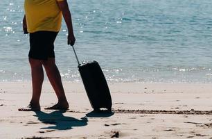 cibo nudo uomo indossare pantaloncini e maglietta trasporto un' nero viaggio valigia su sabbioso spiaggia con turchese mare sfondo, estate vacanze concetto foto