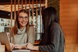 Due giovane attività commerciale donne seduta a tavolo nel bar. ragazza Spettacoli collega informazione su il computer portatile schermo. ragazza utilizzando smartphone, blogging. lavoro di squadra, attività commerciale incontro. foto