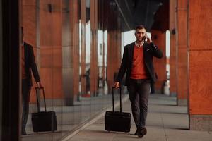 andando per aeroporto terminale. fiducioso uomo d'affari viaggiatore a piedi su città strade e traino il suo valigia potabile caffè e A proposito di su smartphone foto