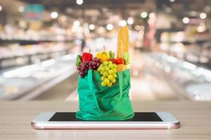 fresco cibo e verdure nel verde shopping Borsa su mobile smartphone su legna tavolo con supermercato corridoio sfocato sfondo drogheria in linea concetto foto