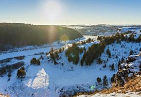 inverno paesaggio con innevato fiume, scogliere con pino foresta e villaggio a d'oro ora foto