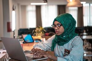 attività commerciale donna indossare un' verde hijab utilizzando il computer portatile nel rilassamento la zona a moderno Aperto Piano avviare ufficio. selettivo messa a fuoco foto