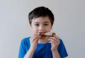 ritratto ragazzo con delizioso viso godere mangiare arrostito pollo bacchetta del tamburo nel il giardino, contento di carino ragazzo mangiare grigliato pollo, salutare cibo per bambini concetto foto