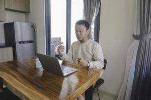 un' giovane asiatico uomo spende il suo tempo a casa seduta nel il cenare camera Lavorando su il suo il computer portatile sentire frustrato foto