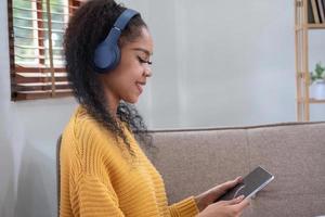 un africano americano donna usi un' computer portatile, smartphone, tavoletta e musica cuffia nel sua casa per relax. foto