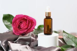 Prodotto per cura della pelle basato su rosa olio, essenziale olio soluzione in un contagocce bottiglia, Marrone pipetta bottiglia e fresco rosa rosa fiore Il prossimo per seta tessile stoffa. foto