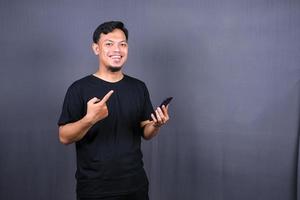 ritratto di giovane asiatico uomo indossare nero maglietta in piedi isolato al di sopra di grigio sfondo, puntamento dito a mobile Telefono foto