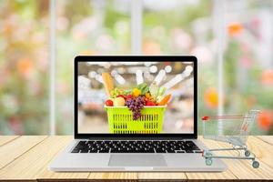 il computer portatile computer e shopping carrello su legna tavolo con finestra e giardino astratto sfocatura sfondo drogheria in linea concetto foto