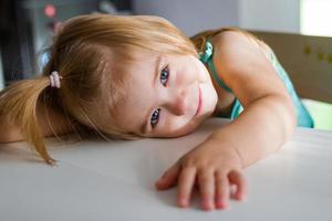 adorabile bambino ragazza con biondo capelli e blu occhi è guardare in il telecamera. soleggiato mattina a casa. foto