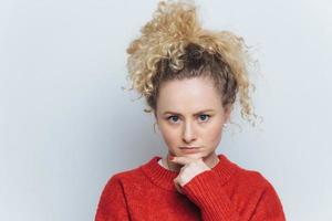 colpo di una donna bionda seria tiene la mano sotto il mento, guarda seriamente la telecamera, isolata su un muro di cemento bianco. lo studente serio ascolta informazioni importanti per l'esame, posa in studio da solo foto