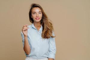 Piacevole donna adorabile con i capelli lunghi, tiene la mano alzata, indossa una camicia alla moda, ha un'espressione da mollusco, sorride piacevolmente, sta in studio su sfondo marrone. persone e concetto di moda foto