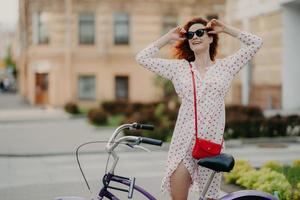 felice donna spensierata pedala in città, posa vicino alla bicicletta, tiene le mani sulle ombre, vestita con un abito bianco, gode dell'ora legale e delle vacanze, concentrata da parte, copia spazio vuoto su sfondo urbano sfocato foto