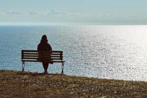 singolo ragazza nel un' nero giacca e cappello seduta su panchina a scogliera a davanti di mare tranquillo, calmo silenzioso posto foto