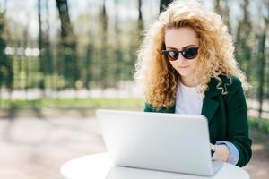 studentessa seria con soffici capelli biondi che indossa una giacca verde e occhiali da sole seduti all'aperto facendo la sua carta di diploma godendo del bel tempo soleggiato. moda, bellezza, concetto di gioventù foto