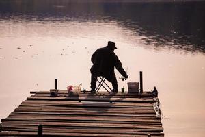 silhouette di un' pescatore a partire dal dietro a seduta su un' di legno molo durante sera pesca foto