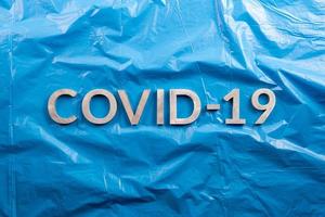 il parola covid-19 di cui con alluminio lettere su spiegazzato blu plastica film sfondo nel piatto posare composizione, direttamente sopra Visualizza foto