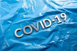 il parola covid-19 di cui con alluminio lettere su spiegazzato blu plastica film sfondo nel piatto posare composizione, diagonale prospettiva foto