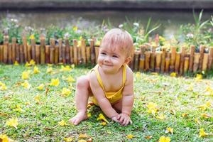 sorridente bambino ragazza con biondo capelli è giocando su erba con giallo fiori su un' soleggiato giorno nel parco. foto