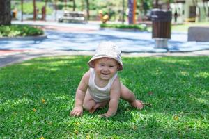 stile di vita ritratto di sorridente bambino ragazza nel bianca capi di abbigliamento e Panama. bambino cerca per strisciare su erba nel parco su un' soleggiato estate giorno. foto