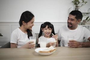 un' salutare asiatico tailandese famiglia, un' poco figlia, e giovane genitori bevanda fresco bianca latte nel bicchiere e pane gioia insieme a un' cenare tavolo nel mattina, benessere nutrizione casa prima colazione pasto stile di vita. foto