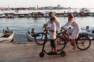 contento famiglia godendo un' bellissimo mattina di il mare insieme, genitori equitazione un' bicicletta e loro figlio equitazione un elettrico scooter. selettivo messa a fuoco foto