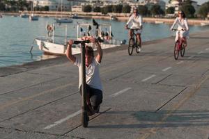 contento famiglia godendo un' bellissimo mattina di il mare insieme, genitori equitazione un' bicicletta e loro figlio equitazione un elettrico scooter. selettivo messa a fuoco foto