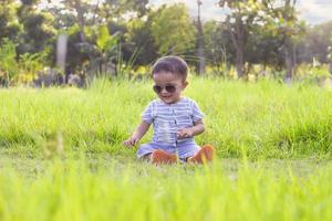 sorridente poco ragazzo ubicazione su erboso campo nel parco, bambino ragazzo con occhiali da sole foto
