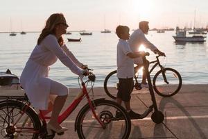 contento famiglia godendo un' bellissimo mattina di il mare insieme, genitori equitazione un' bicicletta e loro figlio equitazione un elettrico scooter. selettivo messa a fuoco foto