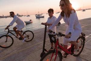 contento famiglia godendo un' bellissimo mattina di il mare insieme, genitori equitazione un' bicicletta e loro figlio equitazione un elettrico scooter. selettivo messa a fuoco foto