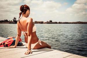 posteriore Visualizza di donna nel costumi da bagno seduta su un' molo. foto