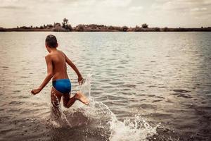 giocoso ragazzo in esecuzione attraverso acqua nel estate giorno. foto