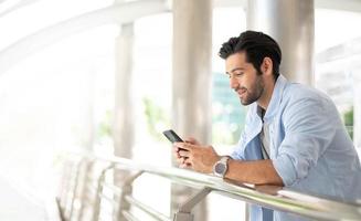 giovane caucasico uomo utilizzando inteligente Telefono e sorridente mentre in attesa il suo amico a il fuori. il uomo Tenere e utilizzando cellulare per ricerca dati e sociale media su Internet. foto