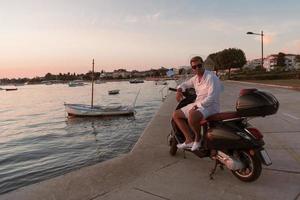 un' moderno anziano uomo d'affari nel casuale Abiti seduta su un' scooter di il mare e gode il Alba. selettivo messa a fuoco foto