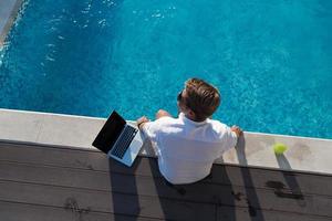 un' moderno anziano uomo gode il piscina mentre Lavorando su il suo il computer portatile Il prossimo per un' moderno lusso Casa. selettivo messa a fuoco foto